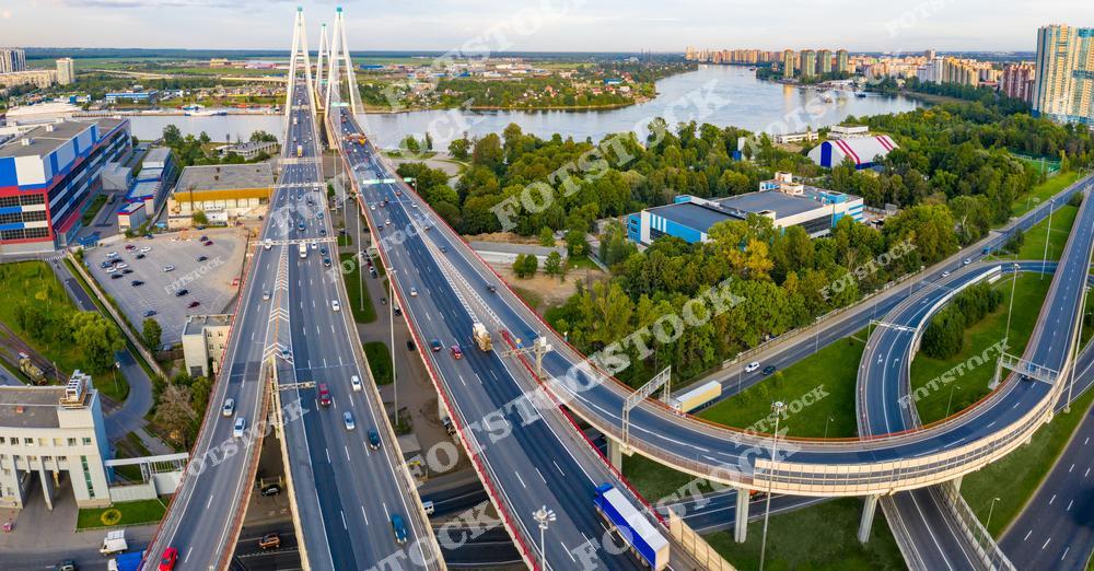 На фотографии изображен Вантовый мост через реку Неву и его транспортные развязки. Видны широкие дороги, соединяющие районы города, и зеленые насаждения, обрамляющие магистраль. На заднем плане расположены жилые кварталы и водные объекты, подчеркивающие гармонию природы и городской инфраструктуры. Снимок выполнен в ясный летний день, что придает яркость и динамичность композиции.